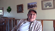 Thumbnail of Monty Hankinson sitting in his house with a wall full of pictures on it and a wooden railing in the background.