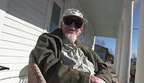 Thumbnail of Bud Lilly sitting on a porch outside a white building in the sun.