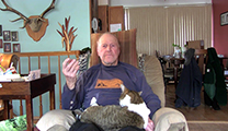 Thumbnail of Tony Schoonen sitting in his livingroom, his diningroom is over one shoulder and a wall with a deer rack over the other shoulder. A cat is sitting on his lap.