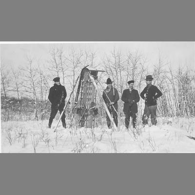 Men Standing in Snow in Front of Bank of Willows and Displaying Sleds and Skis