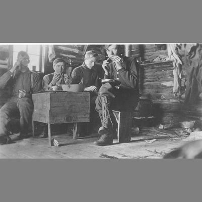 Four Men in the Interior of a Log Cabin Seated at Handmade Wooden Table and Eating a Thanksgiving Meal