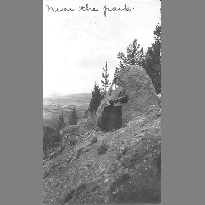 Young Woman Posing with a Rifle in Front of Large Rock on Side of Wooden Slope