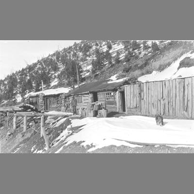 Man Stands in Front of Lengthy Log Structures Built into Slope of Mountain