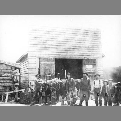 Men and boys posing in front of false-front frame livery stable with team of yoked oxen.