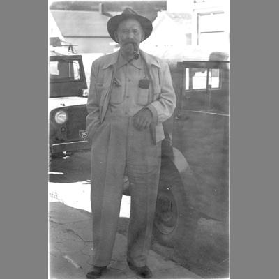 Full View of Joe Redfern Standing in Front of Two Automobiles With a Pipe in His Mouth
