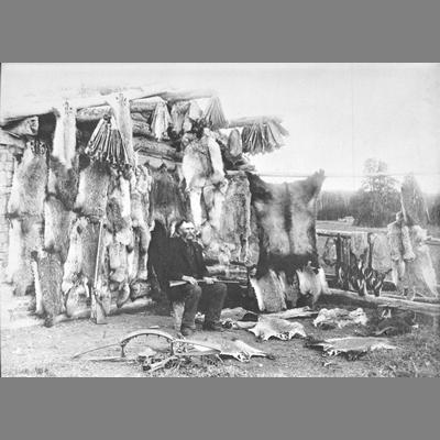 Older Man Seated With Rifle in Front of Log Cabin Decorated with Skins of Dead Animals