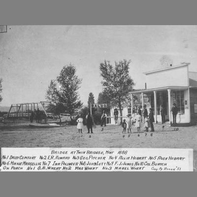 Men, Women, and Children Stand in Dirt Road in Front of Metal Truss Bridge 1888