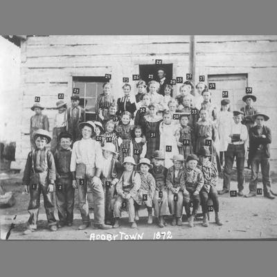 School Children and Teachers Pose In Front of Hewn Log Building - Adobe School, Montana 1872