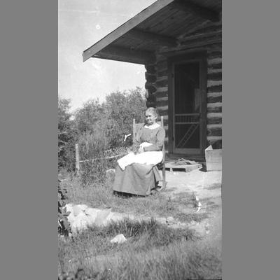 An Old Woman Sits Outside of a Log Cabin on a Rocking Chair