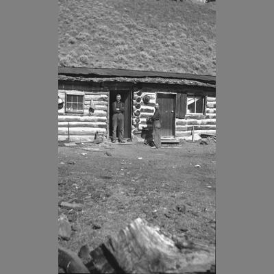 Two Unidentified Men Standing Outside A Log Home