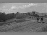 Two unidentified individuals ride horses down a dirt path next to a bridge.