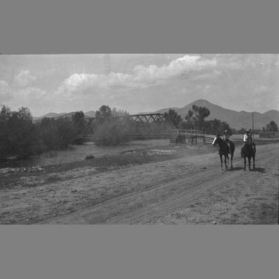 Two unidentified individuals ride horses down a dirt path next to a bridge.