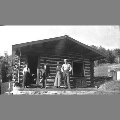 Four unidentified individuals stand on front porch of log home.