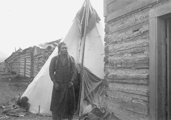 Flathead Indian Man Standing Next to Log Cabin, Tipi Behind Him, Unknown Location