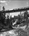 Photo shows freight train with trailer hauling flat cars, box cars, and an open top hopper, crossing bridge near Fish Creek