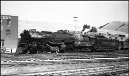 NP 5143, Class Z-8 in front of warehouses at Missoula, with worker crossing in front of locomotive and fireman leaning out of his cab window.