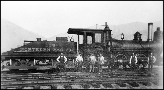 NP 136, a 0-6-0 switcher, at MU Transfer, MT., with five crewmen standing in front of the engine.  This engine was later renumbered to 969.  The tender has a long footboard/walkway the length of the tender hung beneath the tender's side.  