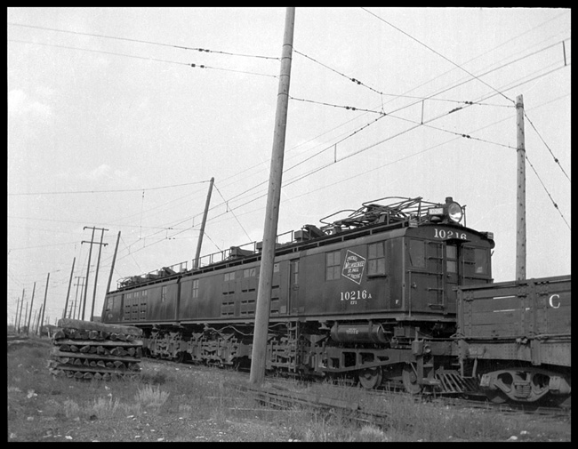 MILW 10216, similar to 10203 which is pictured in Fig. 2159, on page 886 of 1925 Locomotive Cyclopedia by Simmons-Boardman, republished in 1973 by Newton K. Gregg.  The MILW 10203 is reported to be a Class 444-444-E-576-8GE-253A, 3000 Volt direct Current electric locomotive. Locomotive connected to flat car with side boards in stake pockets.