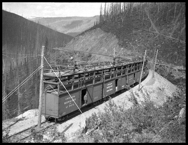 View of Bitterroot Mountains during the electrification of the MILW RR.  Old negative, with no dates or location identified.  Scaffold was built on top of box cars to make a work platform for linemen to work from in installing catinary.