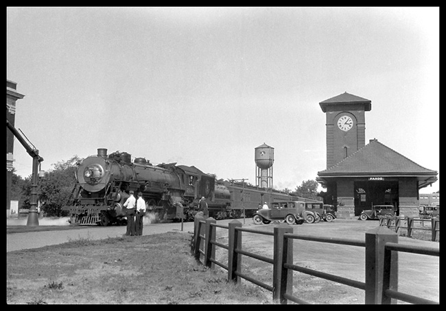 GN Train No. 1 2524 Fargo, ND. GN class P-2  4-8-2  Baldwin 1923  Train at depot  Depot clock  Water column  Unknown onlookers  Several autos of the era Large water tank in background  