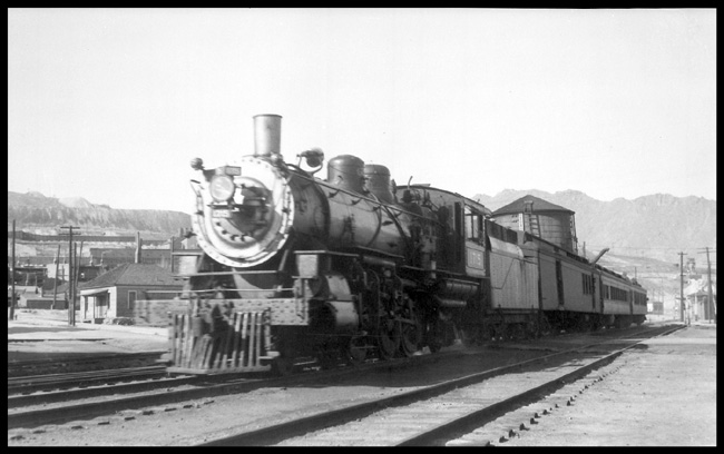 GN #235 1?15 arriving Butte  5/32 GN three car psgr train on unprotected grade crossing, bell ringing  Wood water tank on left partially obscured by train  Depot on right behind train  [backing in?]  LF view