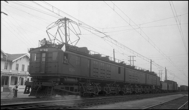 CMSP&P  E36  Three Forks  8/24/40  E36 is a box cab freight motor  It is flying white flags, indicating that it is an extra train  The building in the left background is a hotel, but it appears to have an apparatus in front of it for serving up train orders  This is an unusual shot, taken into the sun from the north side of the tracks, apparently made possible by an overcast day  Most shots of this area are taken from the south