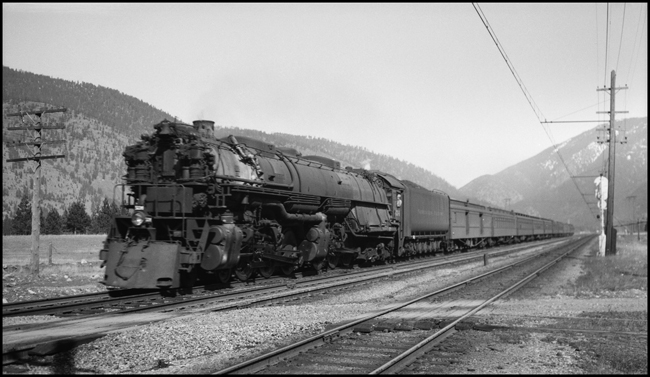 Second No. 1, a section of the westbound North Coast Limited powered by Class Z-8 4-6-6-4 no. 5134, passes the Deer Creek Road grade crossing at high speed--proabably around 80 miles per hour. Engineer Albert A. Melton is at the throttle of no. 5134. His fireman's date is August 25, 1905, and he was promoted to engineer on January 1, 1910. This location is about milepost 115 on the Rocky Mountain Division between Bonner and Missoula. Bonner is located at milepost 113.2 and Missoula is located at milepost 119.4. This was a high speed double track line, and westbound passenger trains typically ran as fast as possible between Bonner and Missoula. Operating rules allowed regular (scheduled) trains to arrive at stations early, but not to leave early. By passing the last station (where time was shown in the Employee's Timetable) prior to a scheduled stop at the highest possible speed, a locomotive engineer could make up a little time if he was running late, or he could arrive a little early at the scheduled stop so as to allow more station dwell time. The track closest to the camera is the electrified line of the Milwaukee Road, which closely parallels the NP line at several points between Garrison and Missoula.
