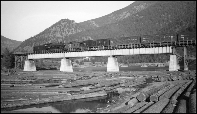 MILW 1220 on bridge over Blackfoot River at Bonner, MT.  River is full of float logs. Locomotive appears to be helping this train.