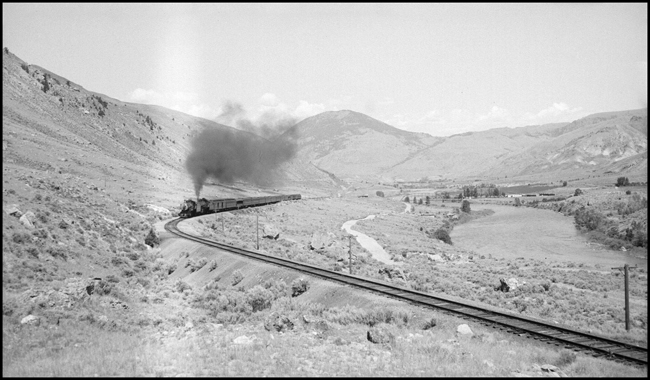 The second engine seems to be working the hardest, as a doubleheader hauls a very large btanchline passenger train toward a curve near the end of its run.  The scene is on the line toward Yellowstone National Park.