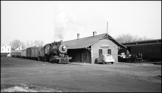 Freight train with NP 328, Class S-10, arriving on one side of Grantsburg Depot while someone is unloading baggage car onto a horse drawn baggage wagon on opposite side of depot.