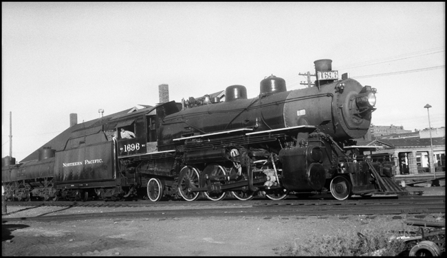 NP 1696, Class W-1 switching tank cars on the north side of the yard office at Livingston, MT.  Industry track to the Sawyers Grocery store in foreground.  Livingston Depot across the tracks from the front of the locomotive.