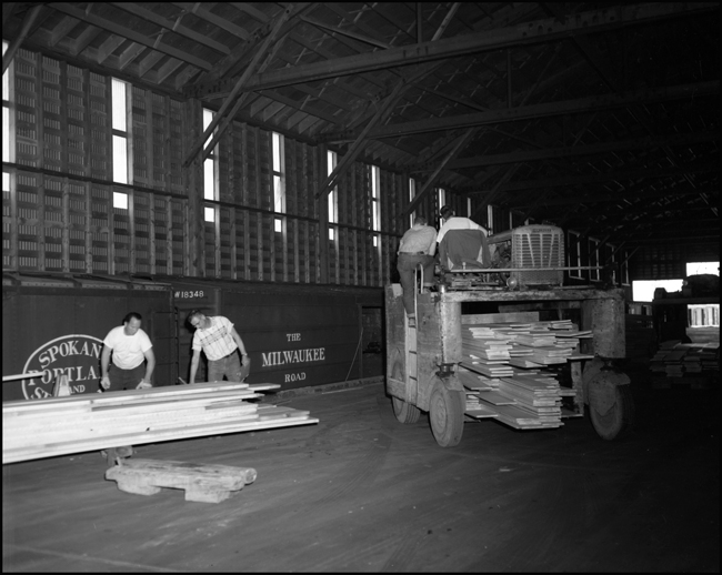 While a Rube Goldburg vehicle brings more lumber for the loading shed a pair of timber graders assess the quality of some new lumber before it is shipped out in forty foot boxcars from a lumber mill near Pablo, MT.
