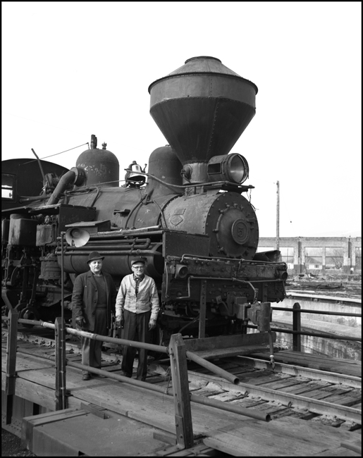 Some local railroadmen take this opportunity to pose with the steam locomotive, a Shay visiting Missoula for minor repair and restoration.  The walls behind the engine suggest that the engine servicing area is going through some post steam era demolition.