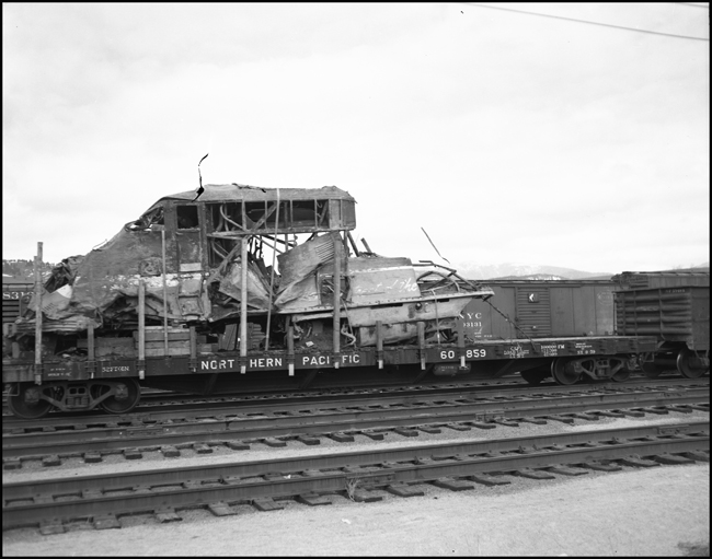 The twisted remains of a part of an F7 cab unit, wrecked in a high speed passenger derailment, are hauled east to the junkyard on a flatcar