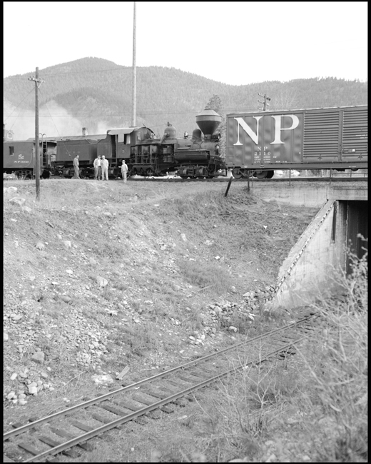 Bearings are running hot on a steam locomotive dead in tow on the Northern Pacific, and a stop has to be made, almost on top of a Milwaukee Road branch line.  Most of the train crew uses this opportunity to pose for a picture