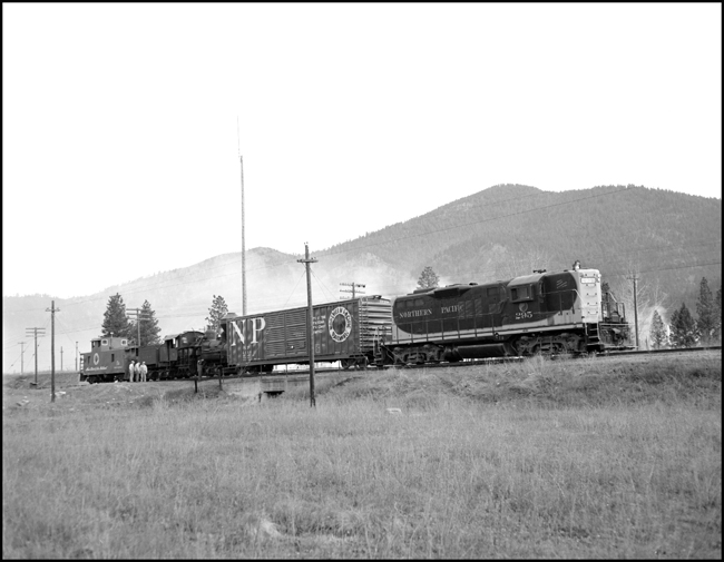 The crew of a special train gathers around its cargo, a steam locomotive in tow that is having a problem with overheated bearings.  They will be under way again in a few minutes.