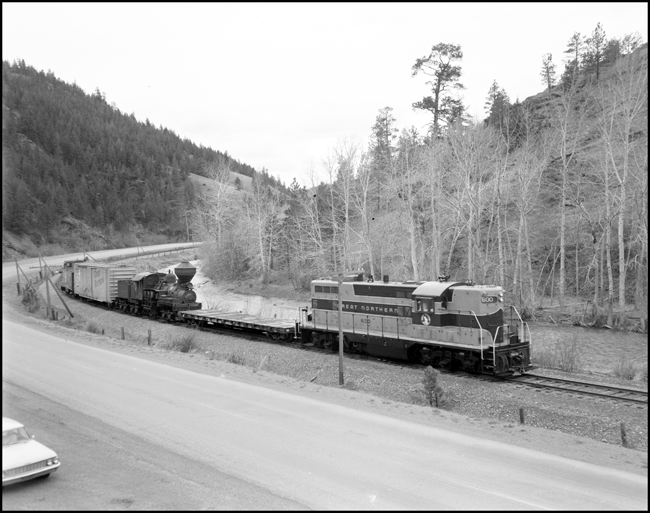 A steamer continues its journey to a new home.  This time the host railroad is the Great Northern,  with a GP-7 diesel, an idler flat, the Shay steamer, a n empty boxcar in the new green colors, and a caboose, head north from Helena.