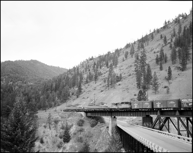 Two Joes and a Geep are going west fast on the Milwaukee, crossing a paved road.  There were 12 Little Joe electrics on the Line and 128 GP-9's, probably the most popular diesel design of the Fifties.
