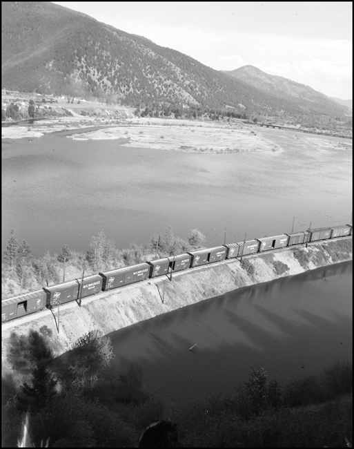 A line of boxcars, empty and with doors open, are probably rolling west for another load of Pacific northwest forest products.  A Milwaukee freight crosses a fill.