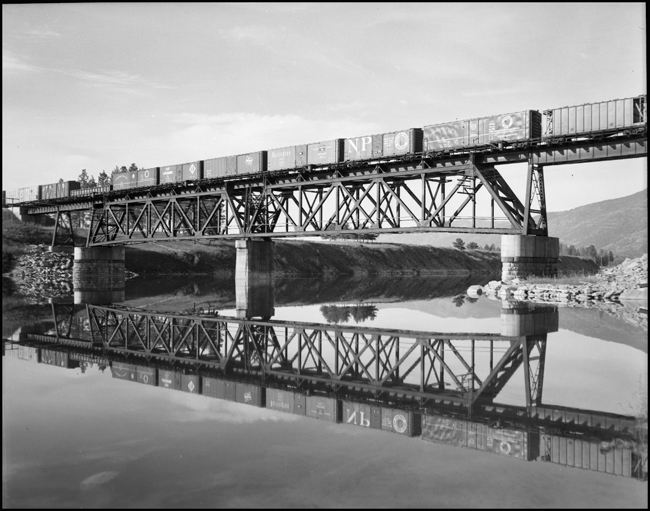 NP XE crossing bridge west of Thompson Falls, and making a mirror image in the water of the train crossing the bridge. 