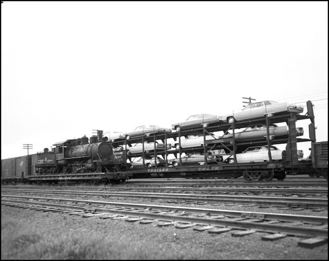 Nez Perce & Idaho 2-6-2 no. 9 on flatcar at Parkwater Yard, Spokane, contrasts with new Cadillacs on eastbound freight.