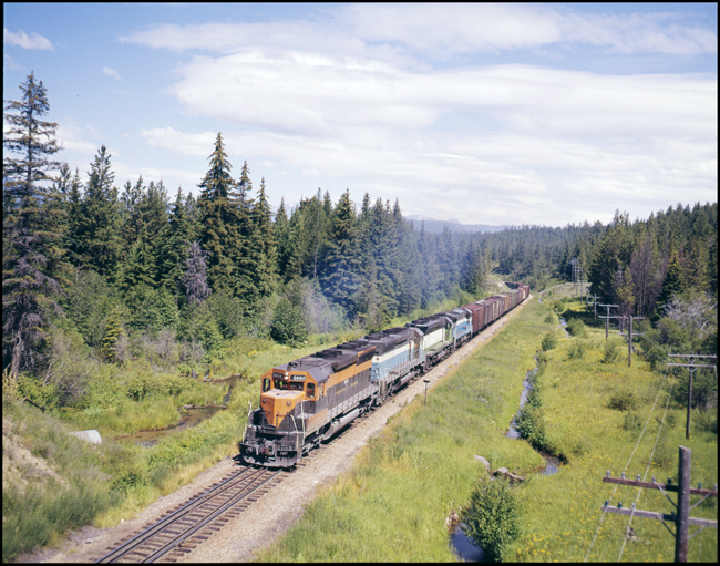 Color picture of BN extra 6446E west of Evaro, MT.  Lead unit in consist is in GN orange and green, second unit is an SD45 in GN sky blue, third unit is an SD45 in CBQ green and white, and fourth unit is a U33C in GN sky blue.