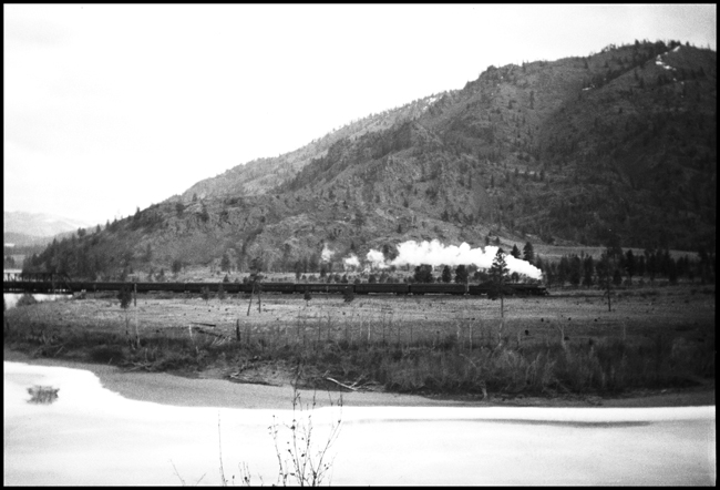 A distance shot of the westbound North Coast Limited, probably in early springtime.  Snow lingers on the hilltops and ice remains in the river.