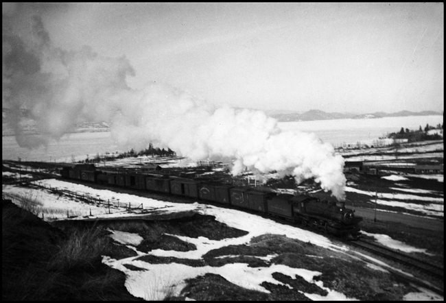 The eastbound local freight makes a dramatic exit from Polson en route to Missoula.  The exhaust from the steam locomotive reminds one of the cotton stuffing exhausts model railroaders would use when photographing their layouts.  No cotton here, just a hard working steam engine on a cold winter day.