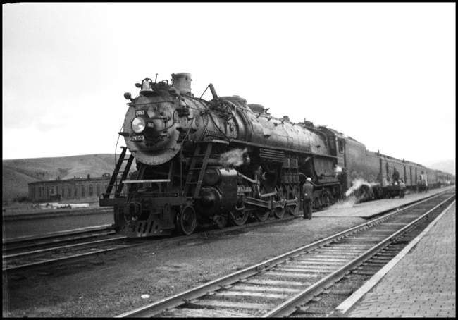 There is action at the doors of the Railway post office and the baggage car as mail and express  are loaded in western Montana.  An A-2 Northern, Baldwin class of 1934, is up front.  The engineer is giving a close look at his power before he starts his run west.
