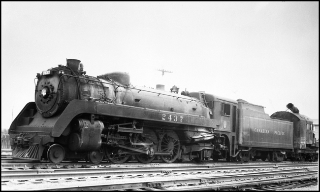 CPR 2437, 4-6-2 locomotive decked out in aerodynamic accessories is connected to a locomotive crane or other piece of work equipment in Winnipeg yard.