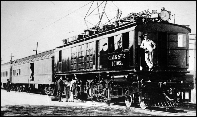 MILW 10105 electric pulling a passenger train, with crew members on platform and on front pilot of locomotive.
