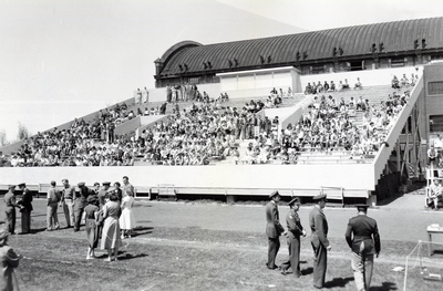 Spectators in the bleachers and people in uniform stand on the field.