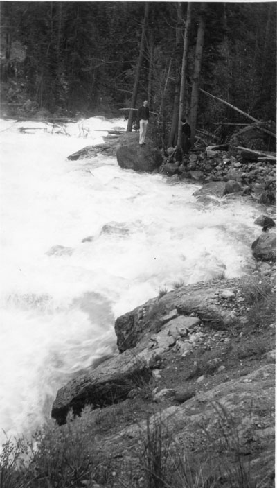 Two people standing along a river, taken from down stream