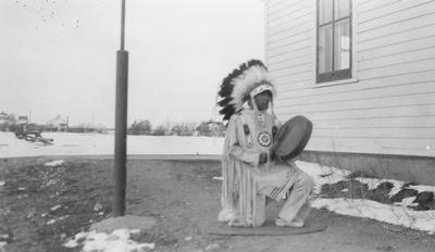 Photo of a young man in ceremonial dress kneeling on a rug and playing a drum, Browning, Montana, 1933